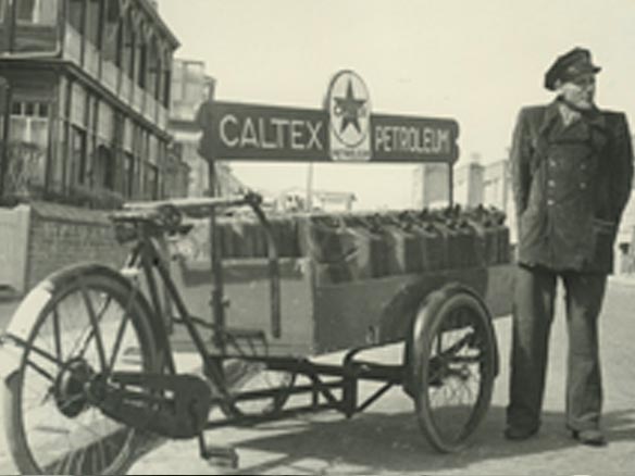 Black and white photo of a man in uniform standing beside a cart with Caltex fuel cans