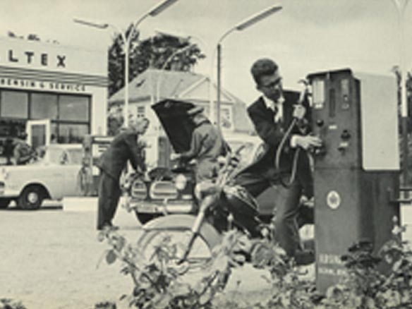 Black and white photo of a man fuelling up and mechanics repairing a car 