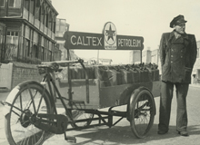 Black and white photo of a man in uniform standing beside a cart with Caltex fuel cans